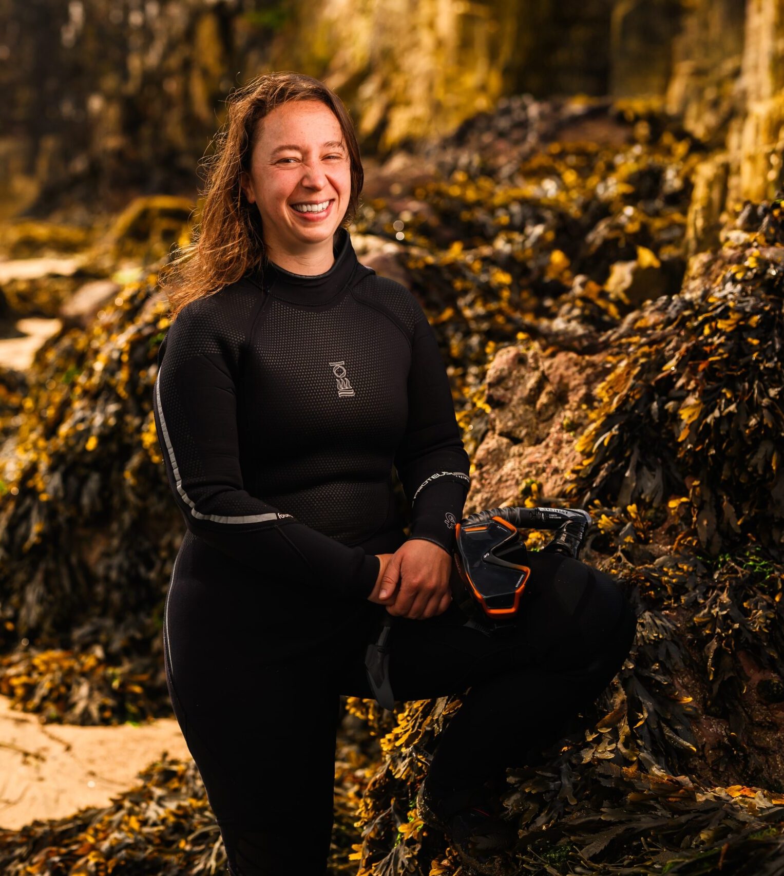Nora Salland smiling on rocky shore in a wetsuit holding mask and snorkel