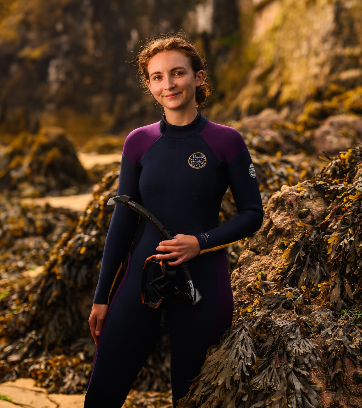 Sophie Corrigan smiling in a wetsuit on the rocky shore surrounded by seaweeds