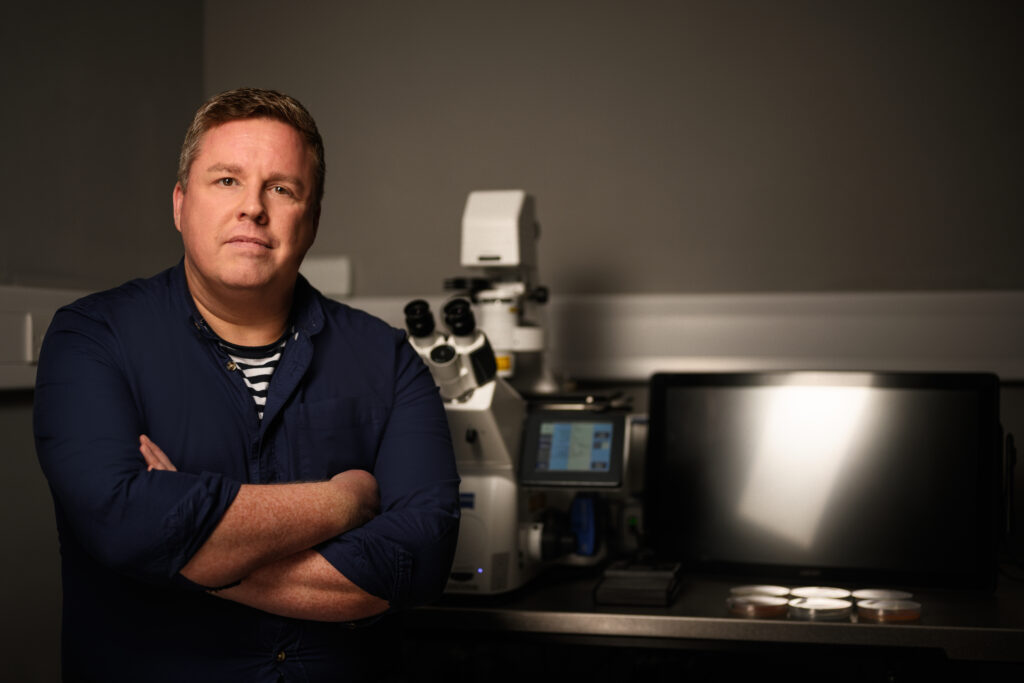 Dr Michael Cunliffe standing with arms folded next to a microscope