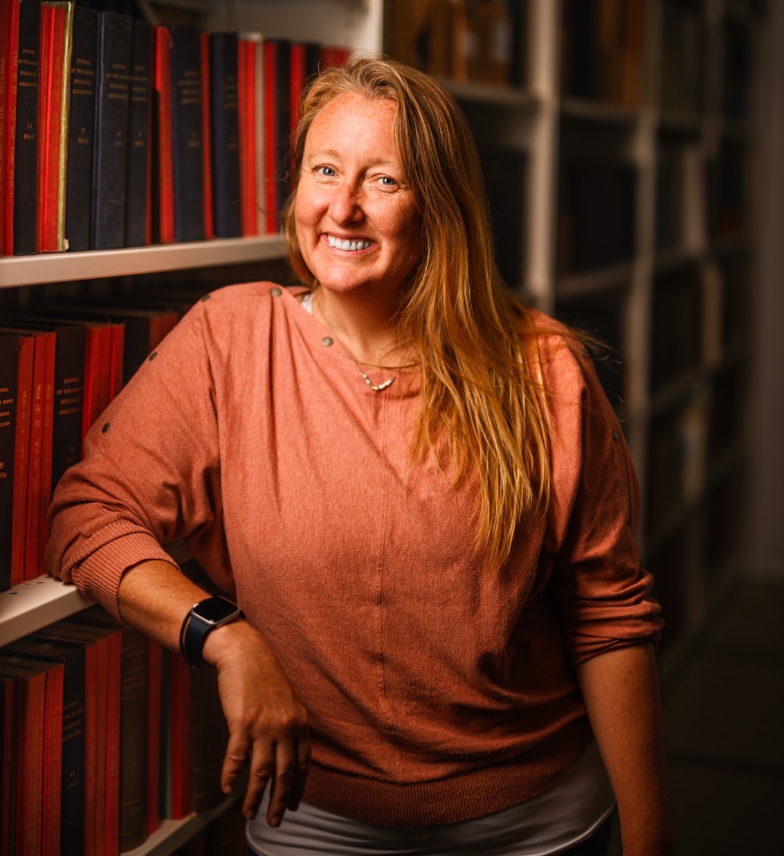 DASSH Data Manager Charlotte (Charlie) Gough leaning against a bookcase in the library.