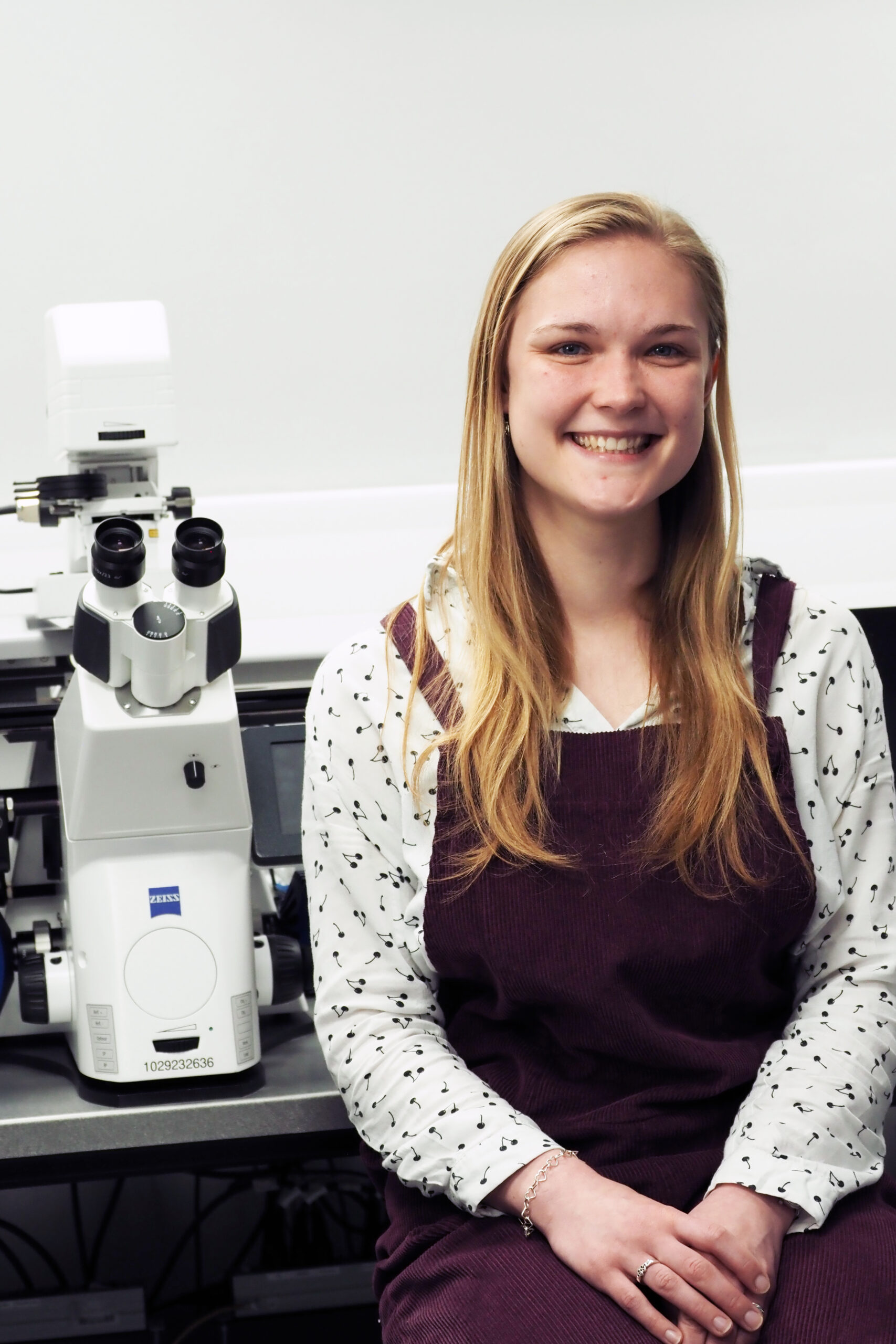 PhD student Beth Tindal-Jones smiling sat next to a microscope