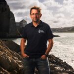 Dr Dan Smale, a man with brown hair in a navy polo shirt and jeans, standing in front of a coastline and sea.
