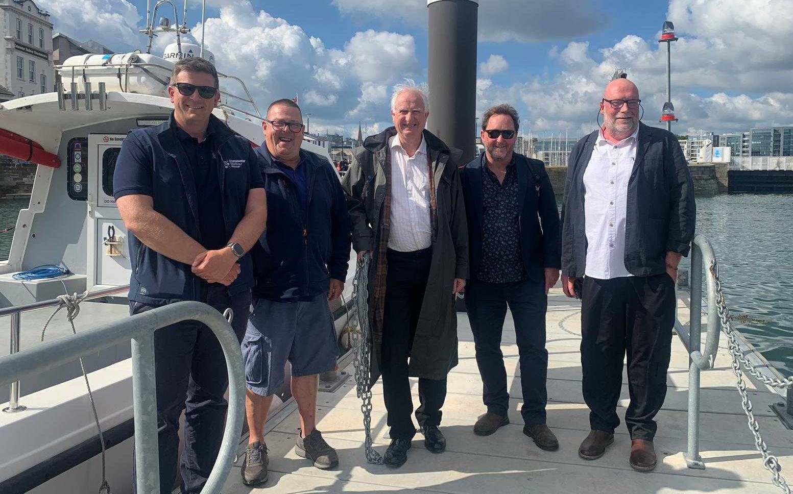 A group of five men stand on a jetty in a marina, a white fishing boat partially visible to the left.