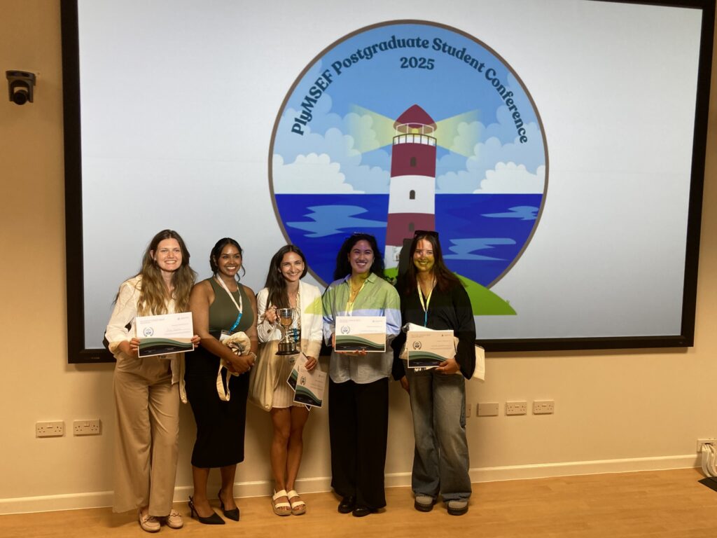 A group of 5 smiling women holding award certificates, the middle figure holding a silver trophy, stand in front of a large poster with a red and white lighthouse on it.