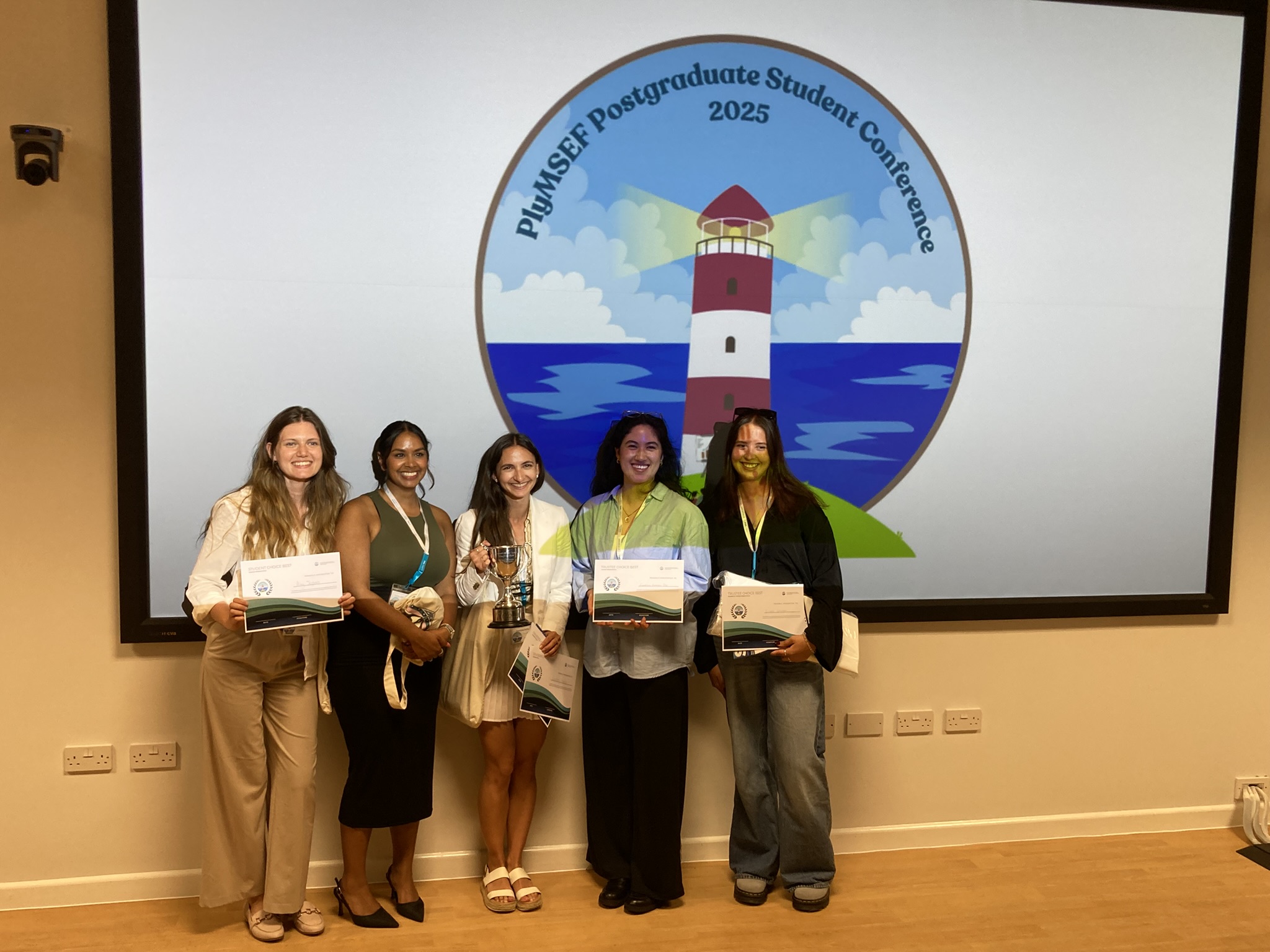6 young women hold up certificates in front of a large round illustration of Smeaton's Tower, a read and white lighthouse.