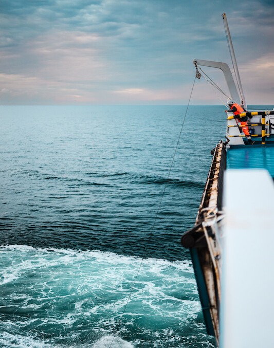 The side of a large boat travelling on the ocean, a man in orange safety gear looks over the side at a rope going from a crane into the water.
