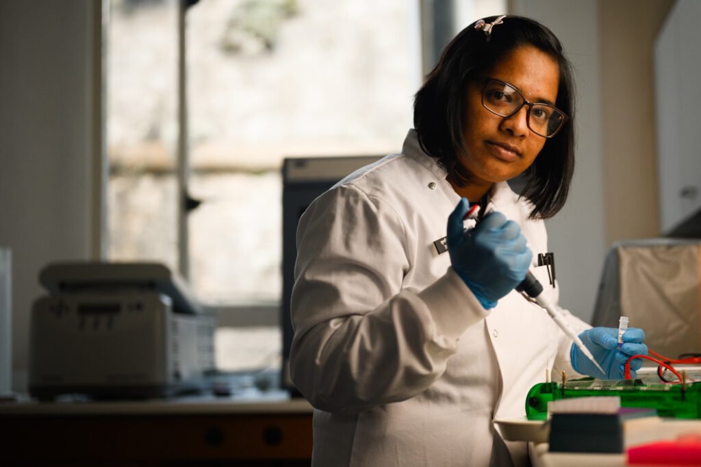 Scientist with dark bobbed hair abd glasse looks solemnly at the camera, in a white coat and holding a pipette.