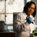 Scientist with dark bobbed hair abd glasse looks solemnly at the camera, in a white coat and holding a pipette.