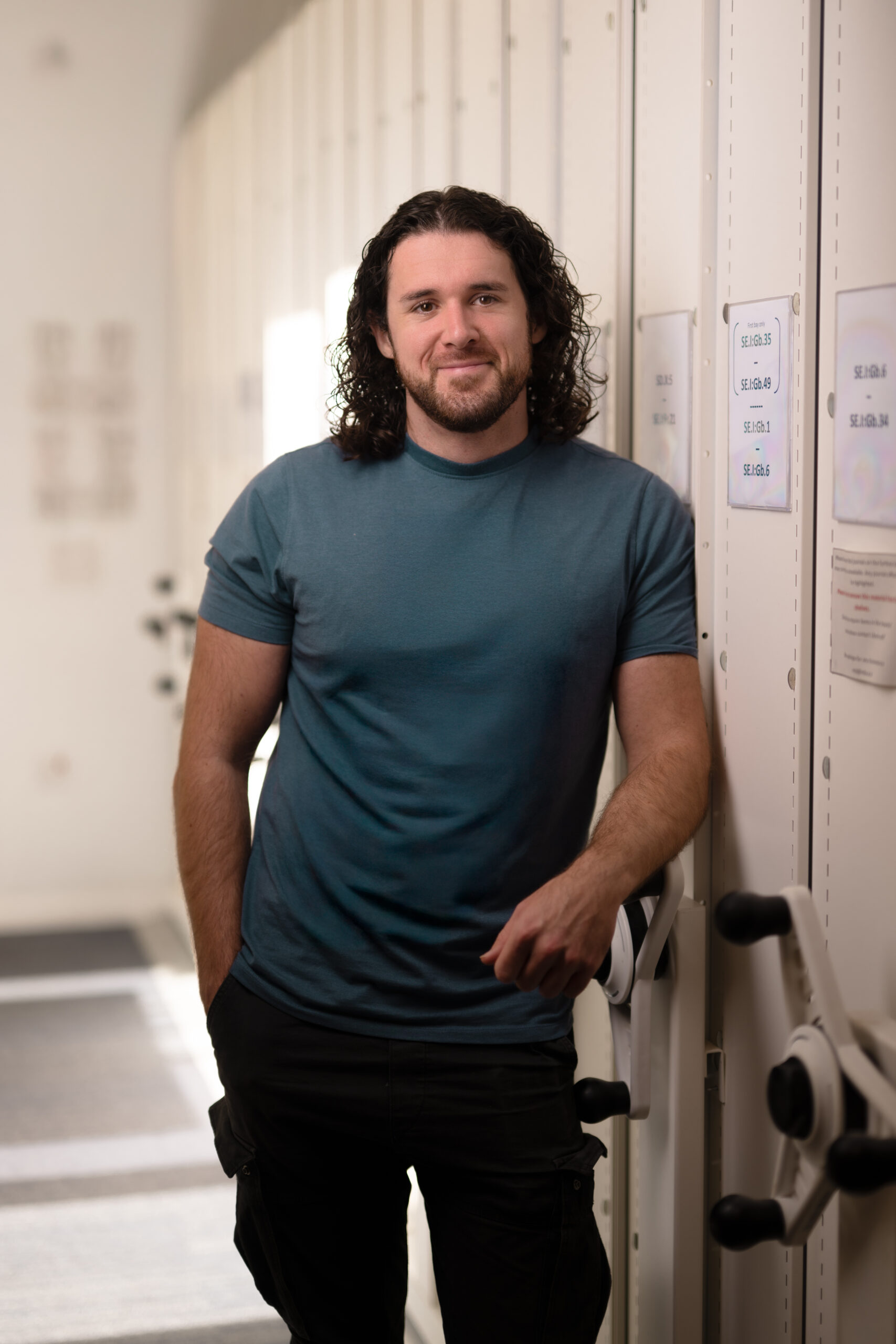 Owen Harris with black shoulder length curly hair, in a green t-shirt, leaning on a library wall.