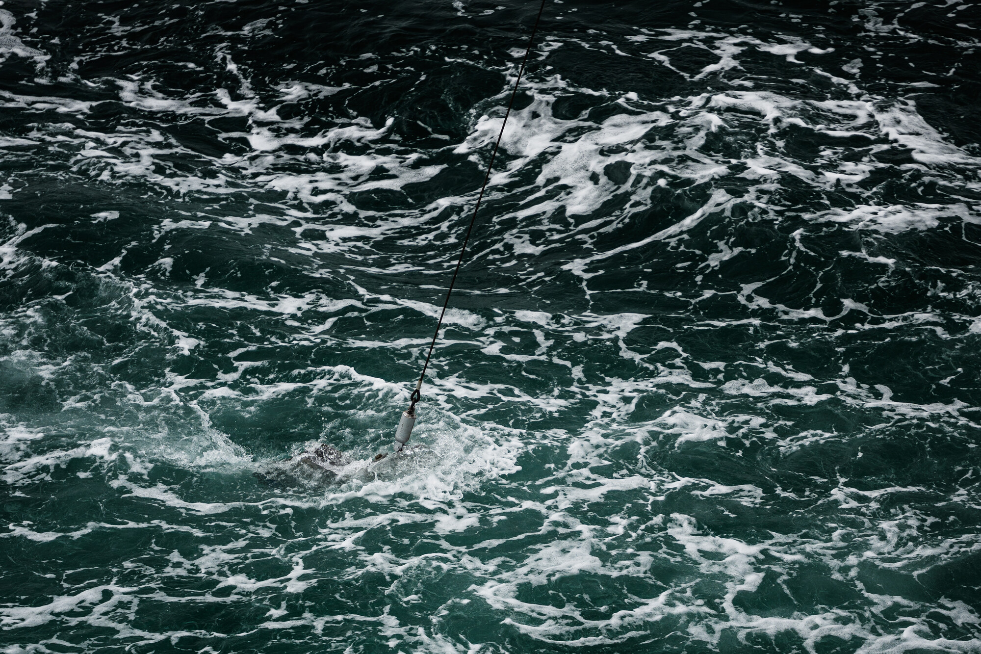 Churning seas, dark with white tips of waves, small grey metal object trailing from steel rope in the cemtre.
