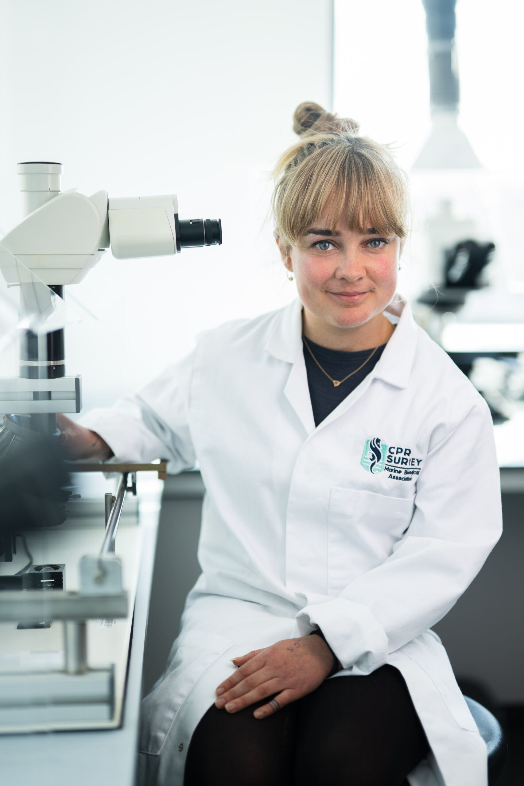 Jasmine Heber-Percy, young woman in white lab coat and blonde tied-back hair with a fridge, sitting next to a microscope on a lab bench.