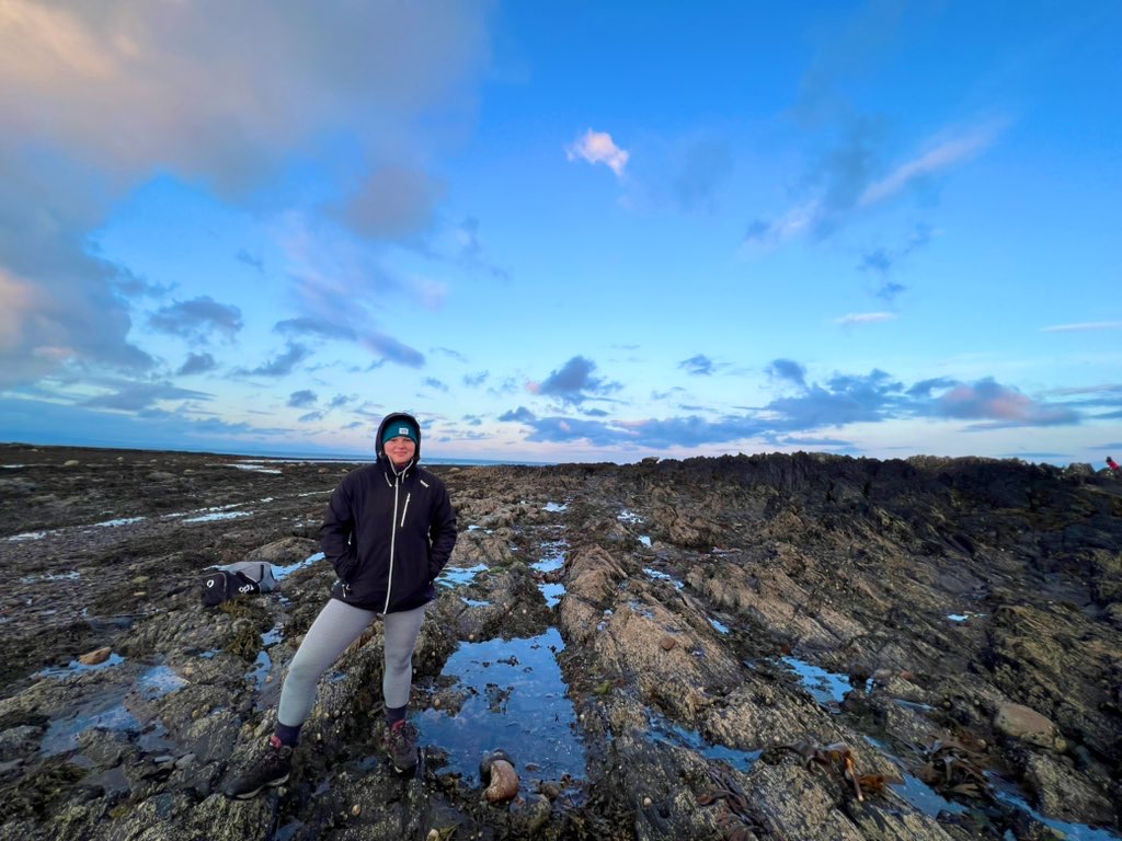 Young woman in casual clothes stands smiling on the rocky foreshore with a blue sky and wispy clouds above.