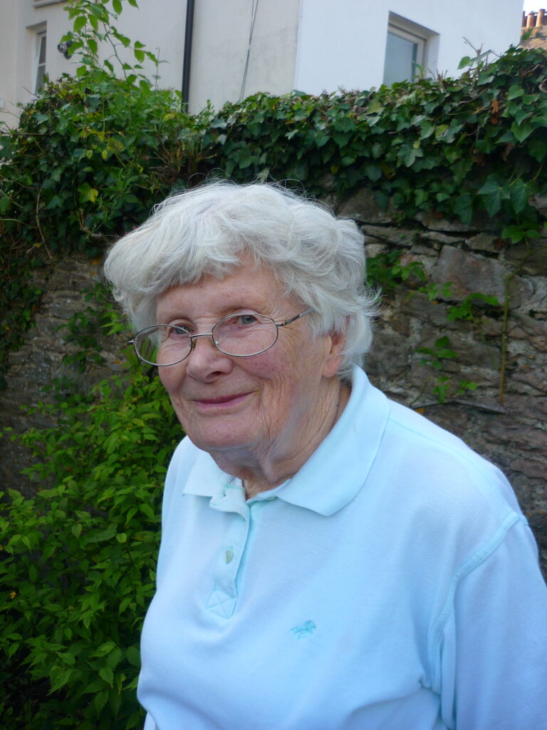 Eve Southward, an elderly lady with white hair and glasses, smiling, in her garden.