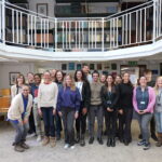 A smiling group of people poses in the National Marine Biological Library under a mezzanine floor with white railings and shelves full of books.