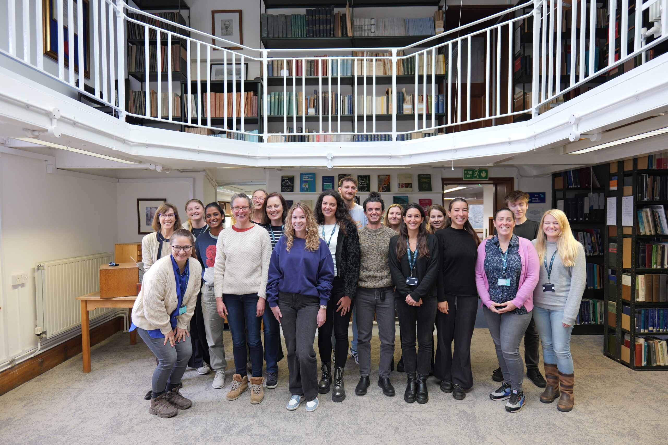 A smiling group of people poses in the National Marine Biological Library under a mezzanine floor with white railings and shelves full of books.