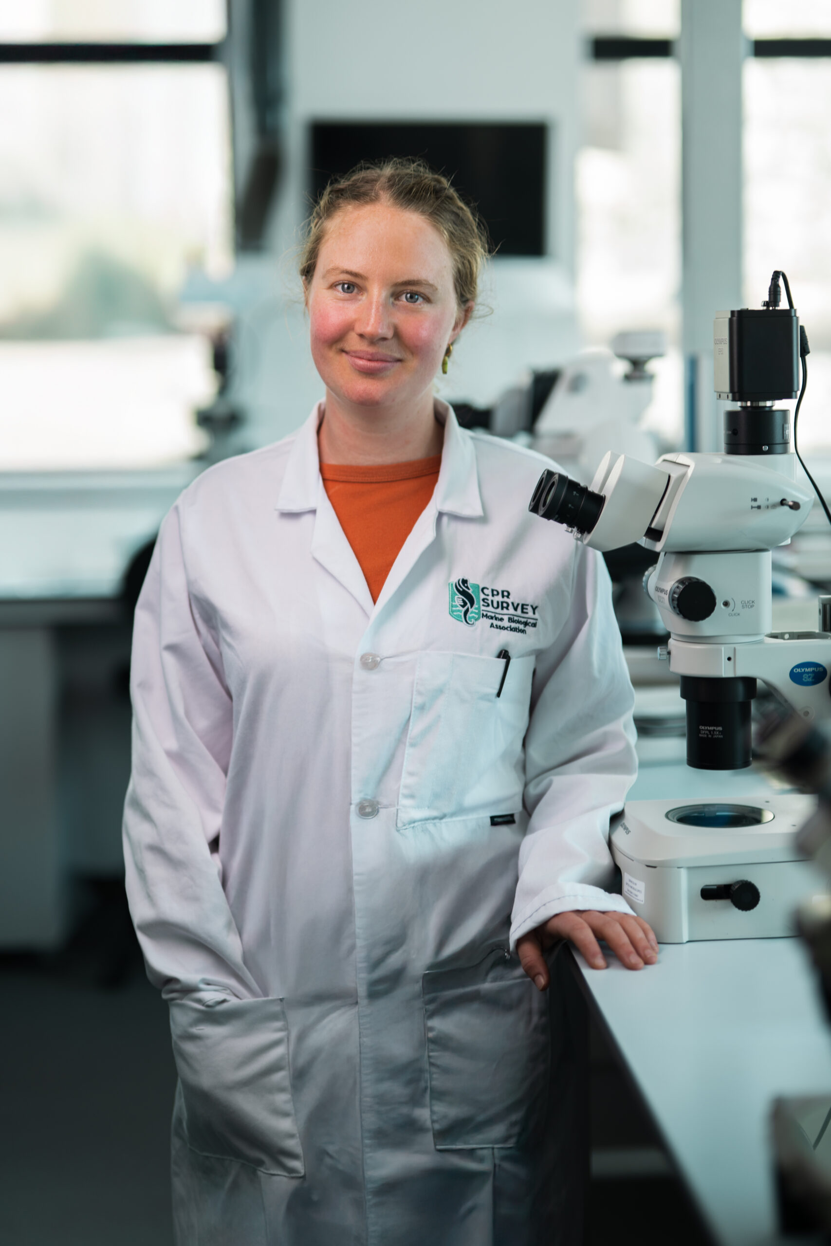 Isabelle Emery, young woman in white lab coat with blond hair tied back, one hand on a lab bench.