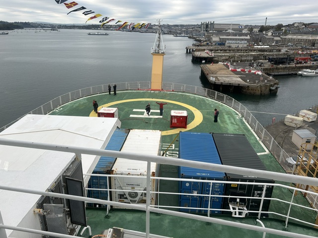 The large bow of a ship facing toward the sea and Devonport Dockyard, the H for helipad on the platform and a person standing on it with