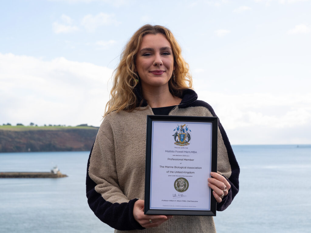 Young woman with wavy long blonde hair, in a brown and black fleece, stands holding a framed certificate, with the sea and headland in the background.