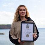 Young woman with wavy long blonde hair, in a brown and black fleece, stands holding a framed certificate, with the sea and headland in the background.