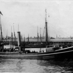 Old black and white photo of the MBA Research Vessel Huxley, a long low boat with a steam chimney, floating on the sea in a harbour with a long low building and other boat masts in the background.