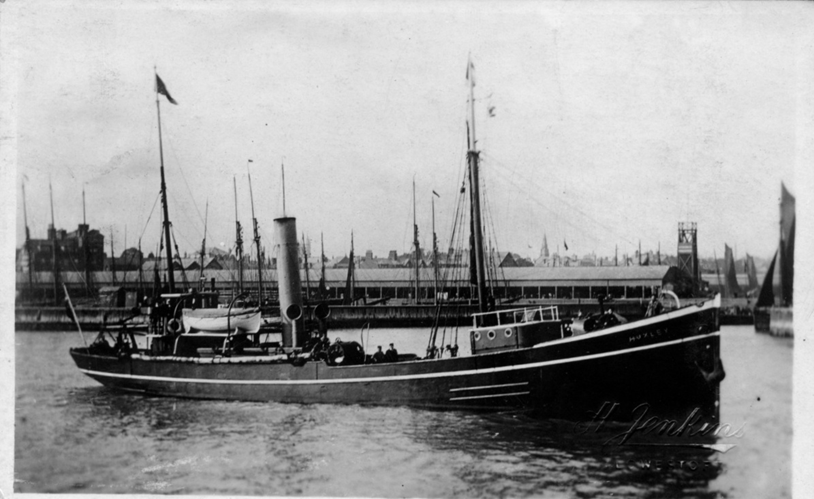 Old black and white photo of the MBA Research Vessel Huxley, a long low boat with a steam chimney, floating on the sea in a harbour with a long low building and other boat masts in the background.