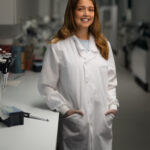 Young woman with long blonde hair poses in a lab net to a bench, in a white lab coat with her hands in her pockets. A pipette is visible on the white lab bench.
