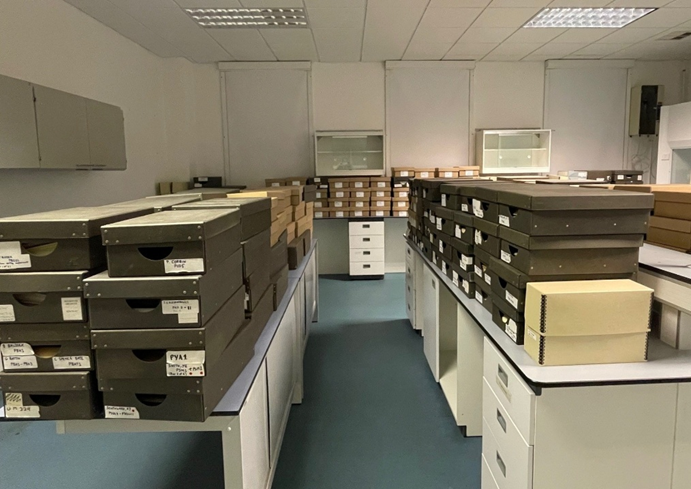 A room with white walls and ceiling and a blue-grey solid floor, with drawers and shelving units, on top of which are dark brown, light brown, and cream boxes.