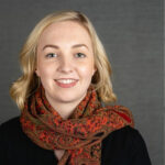 Headshot of a woman with blonde hair in a bob style, smiling at the camera, a red patterned scarf around her neck.