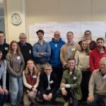 Group photo of adults of different ages in a large room with a clock in the background.