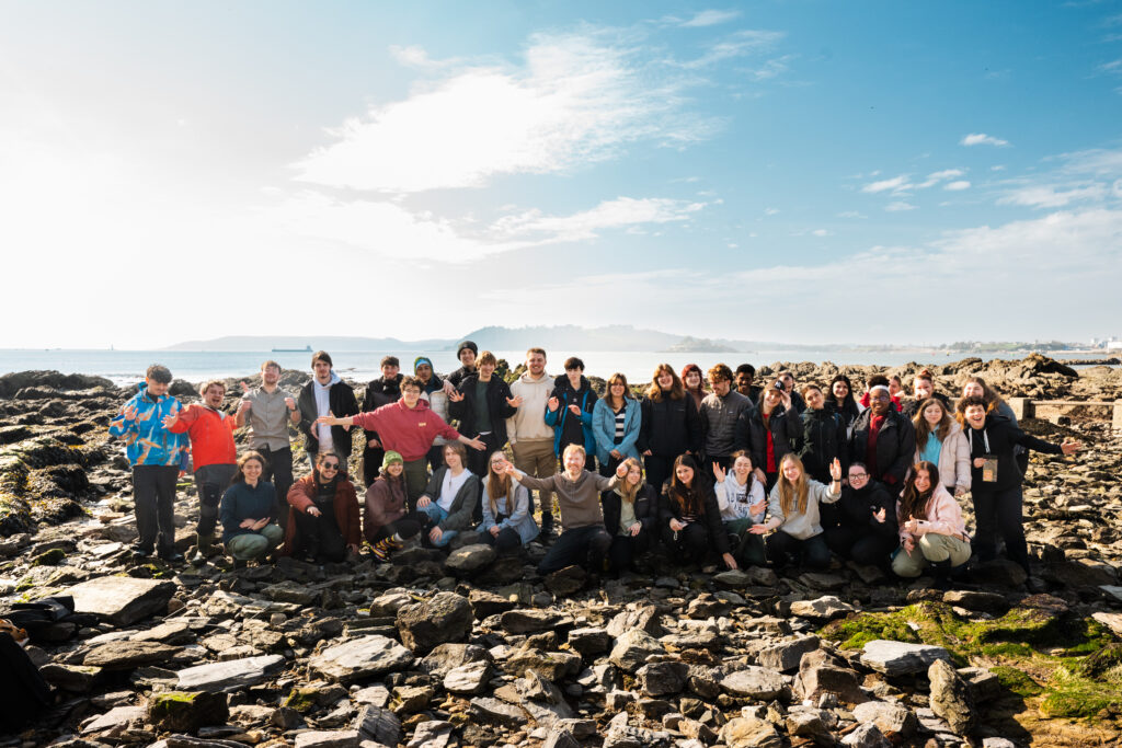 Large group of young people pose for a photo on the rocky shore, with the waters of Plymouth Sound and further land hazily visible in the background.