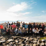 Large group of young people pose for a photo on the rocky shore, with the waters of Plymouth Sound and further land hazily visible in the background.