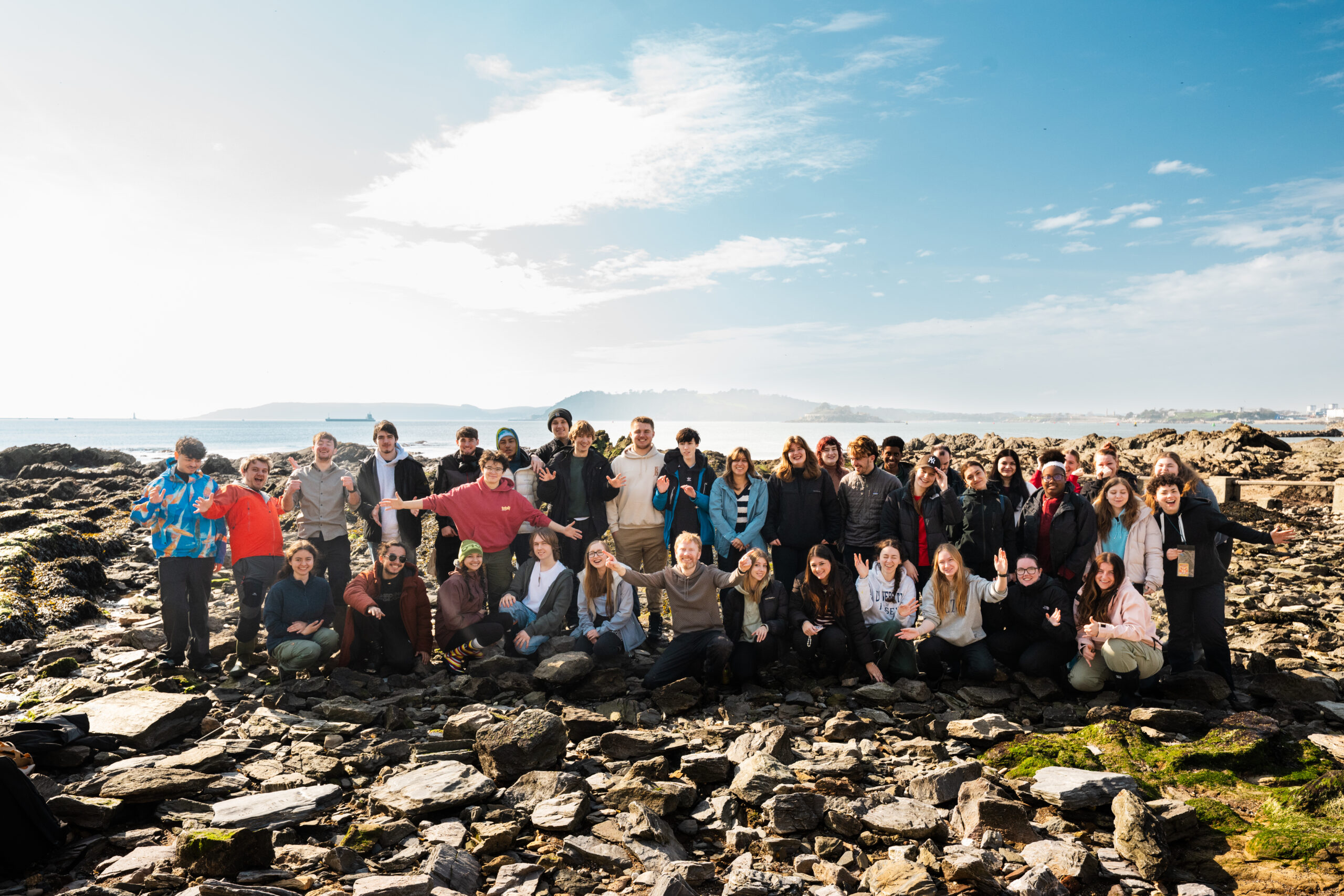 Large group of young people pose for a photo on the rocky shore, with the waters of Plymouth Sound and further land hazily visible in the background.