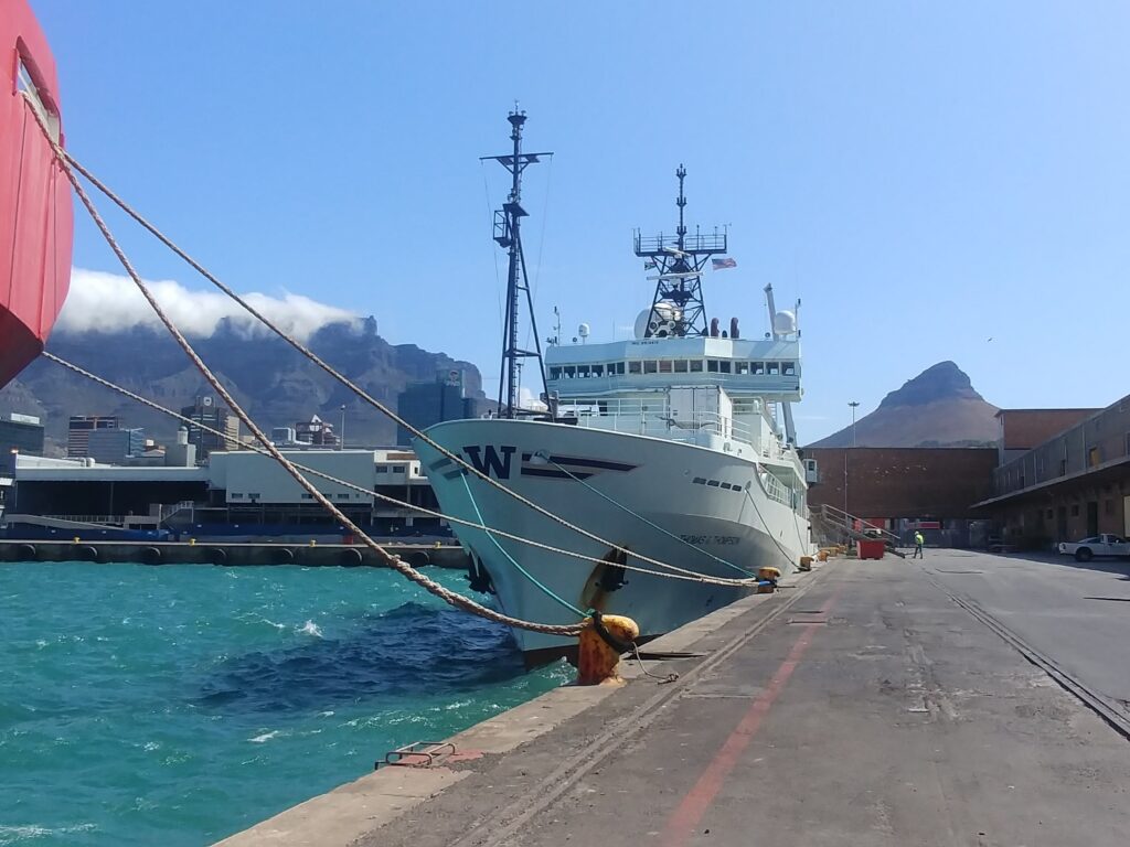 RV Thomas G. Thompson research vessel berthed at Cape Town, prior to departure. Large research vessel by dock, on turquoise sea, with buildings low on the horizon and mountains rising in the background topped by clouds. c. Daniela Sturm.