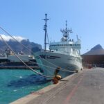 RV Thomas G. Thompson research vessel berthed at Cape Town, prior to departure. Large research vessel by dock, on turquoise sea, with buildings low on the horizon and mountains rising in the background topped by clouds. c. Daniela Sturm.