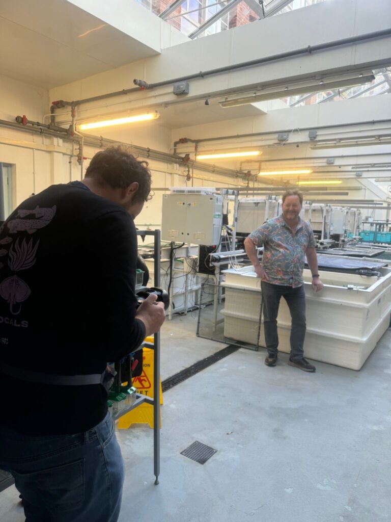 Man with back to the camera filming Dr Bryce Stewart in sea creature-themed short sleeved shirt and jeans, one hand resting next to him on a tank in the MBA's research Aquarium.