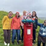 A group of smiling women standing behind a red postbox, topped with a beautiful crocheted postbox topper depicting marine life.