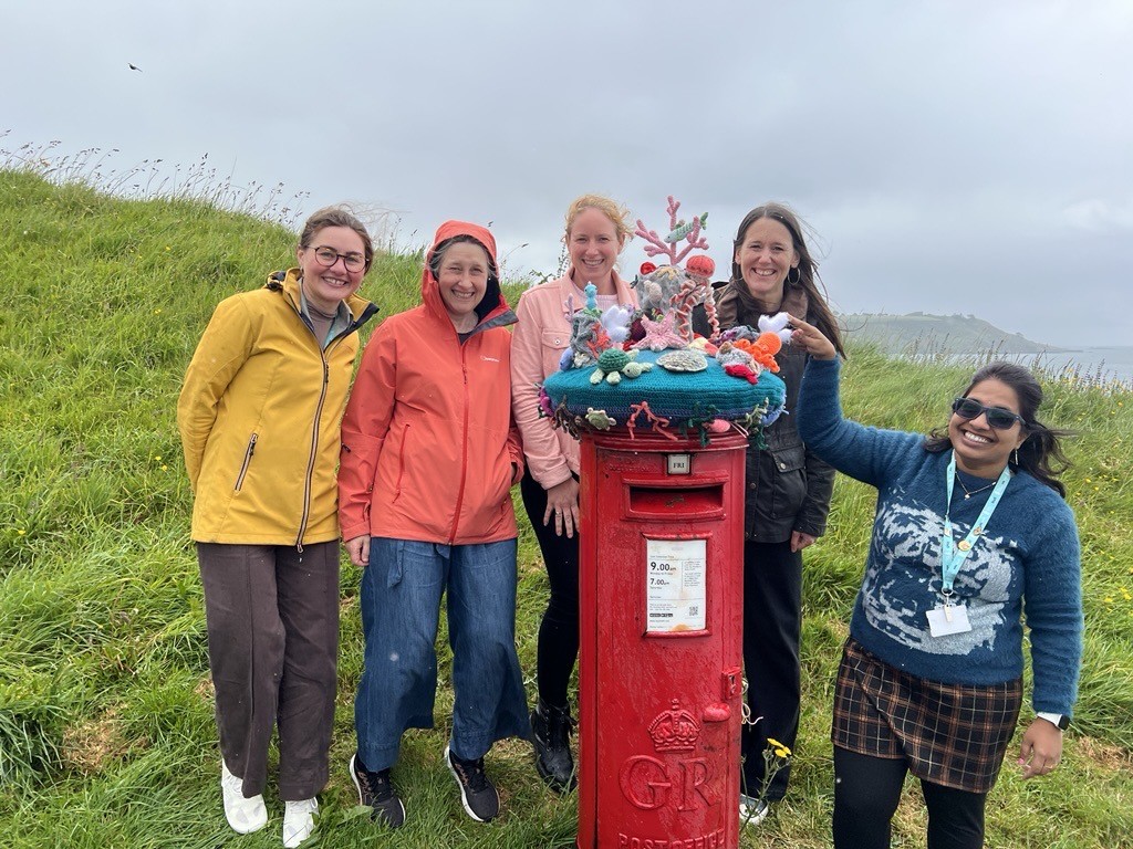 A group of smiling women standing behind a red postbox, topped with a beautiful crocheted postbox topper depicting marine life.
