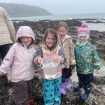 Four little girls standing on a rainy rocky shore beam at the camera as one of them proudly holds up a bucket containing their find, a small crab.