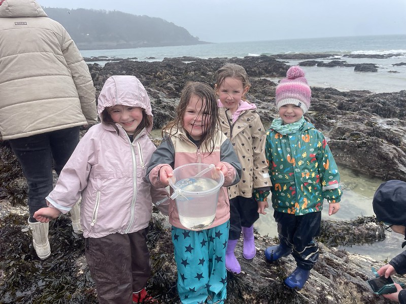 Four little girls standing on a rainy rocky shore beam at the camera as one of them proudly holds up a bucket containing their find, a small crab.
