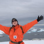 Image of Rod Downie, in a bright orange coat with a WWF logo on it. He is wearing sunglasses and a black beanie hat, and his arms are outstretched, his hands in black gloves. He is smiling and the background is a polar seascape with ice and icebergs visible in the background.