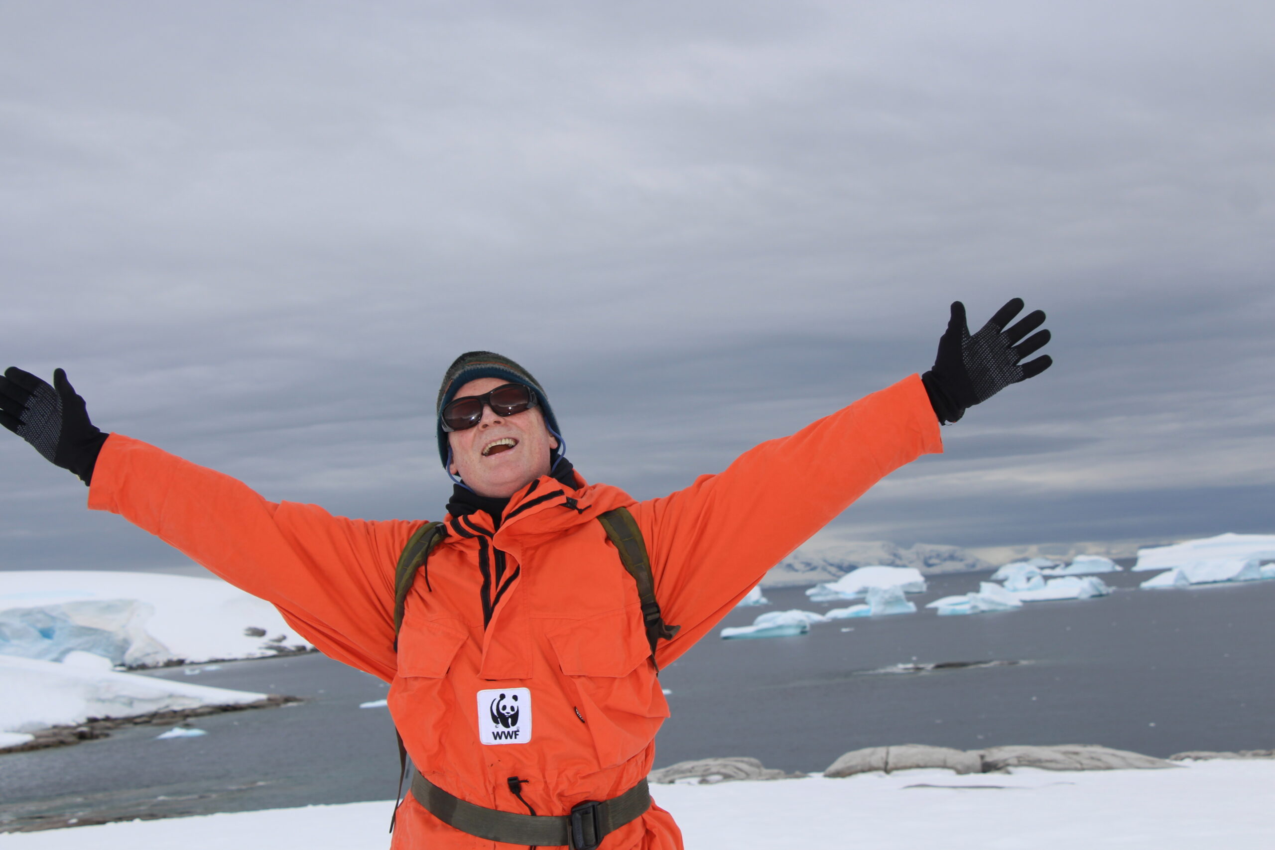 Image of Rod Downie, in a bright orange coat with a WWF logo on it. He is wearing sunglasses and a black beanie hat, and his arms are outstretched, his hands in black gloves. He is smiling and the background is a polar seascape with ice and icebergs visible in the background.
