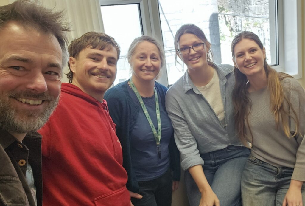 A row of five people in a selfie in front of a window indoors, including left to right and older and younger man, an older lady and two young women, all smiling and dressed in casual clothes.