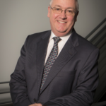 Professor Thomas Miller smiles at the camera, a distinguished gentleman with thinning grey hair and glasses, smiling at the camera, in dark suit jacket, shirt and tie.
