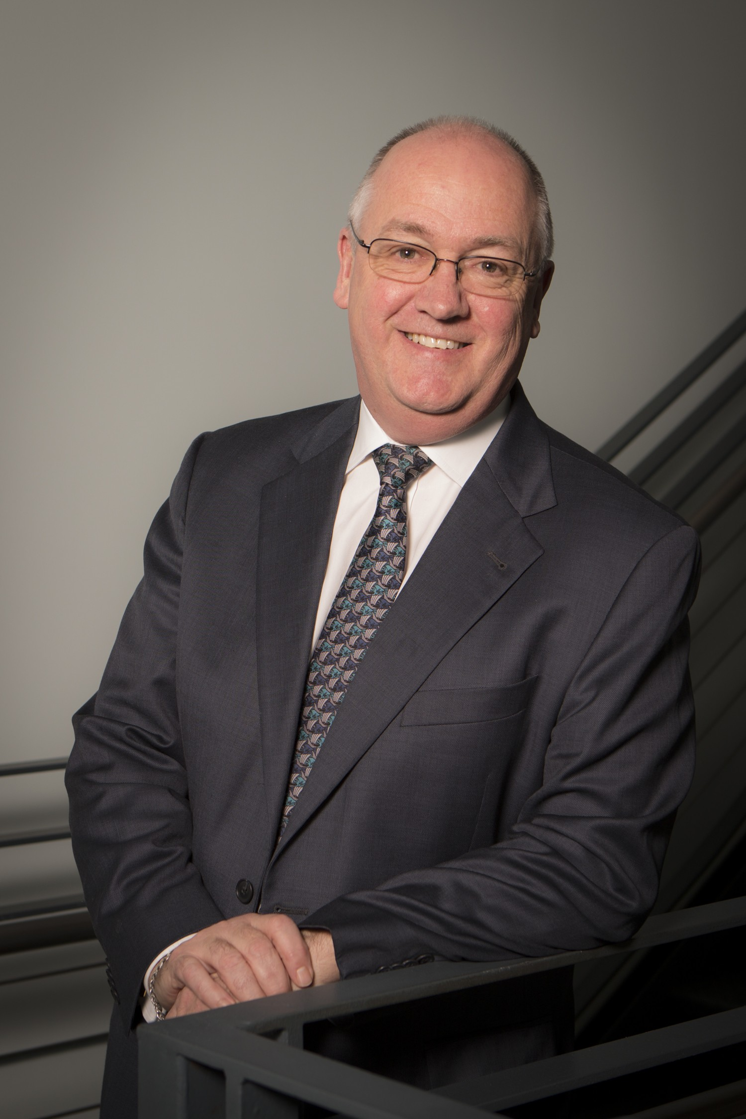Professor Thomas Miller smiles at the camera, a distinguished gentleman with thinning grey hair and glasses, smiling at the camera, in dark suit jacket, shirt and tie.