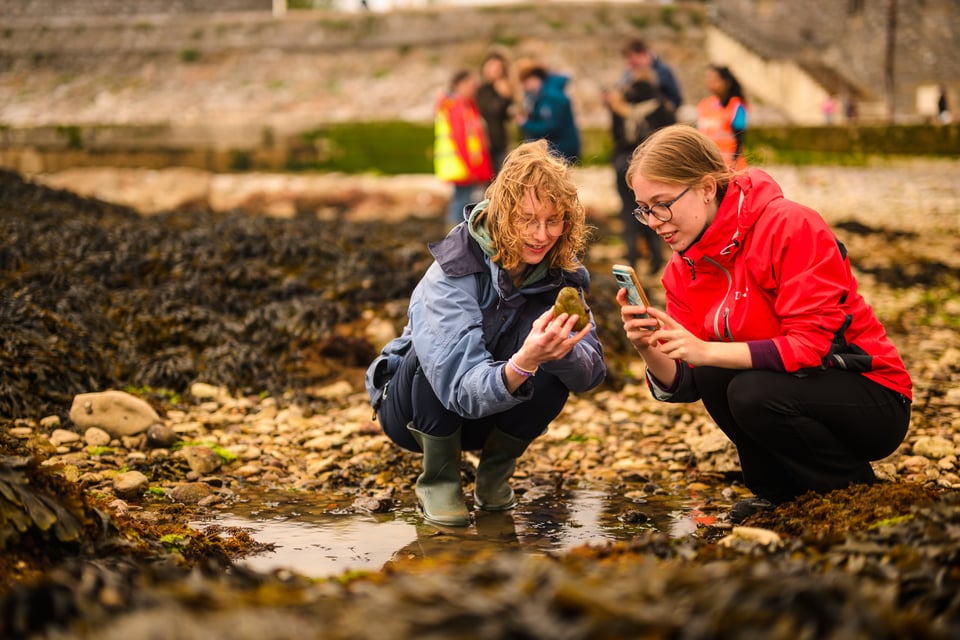 Two young women with blonde hair, in raincoats and wellies, crouch down on the rocky shore, one holding a rock and the other photographing it with her mobile phone. Blurry figures can be seen standing in the background.