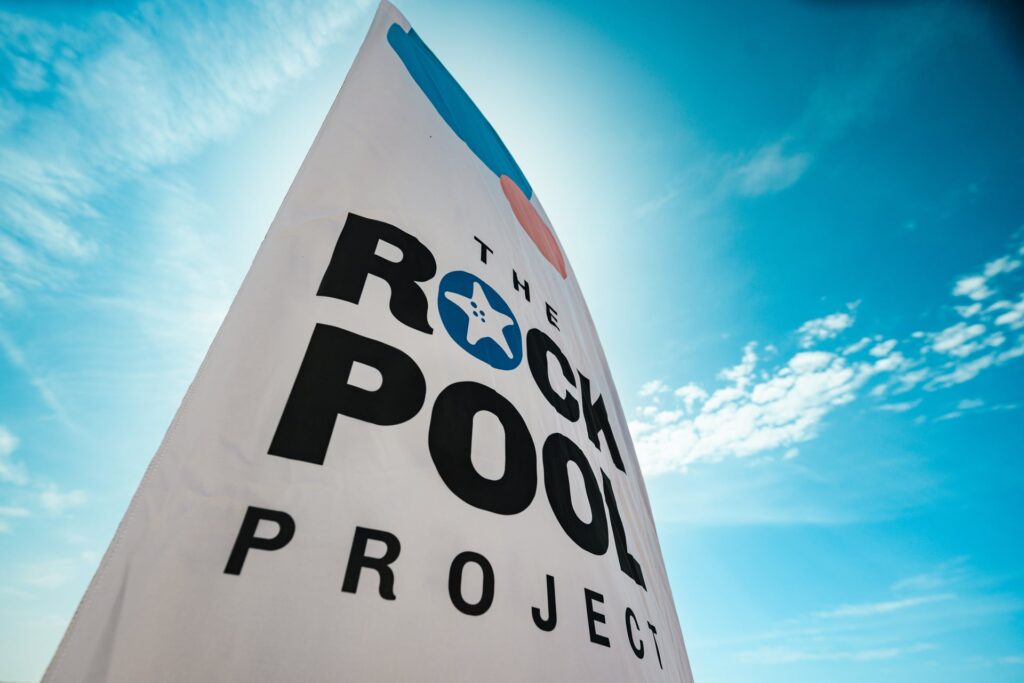 Vertical banner with the words The Rock Pool Project on it, with a sunny blue sky and little clouds behind.