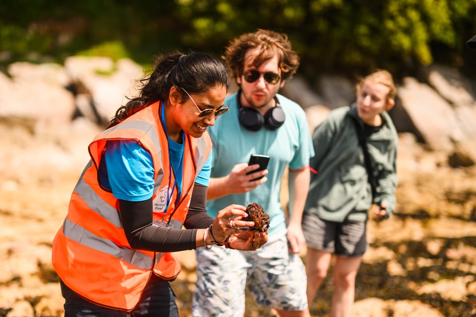 A young woman with dark long hair in a ponytail hair wears sunglasses and a bright orange tabard over a long sleeved t shirt. She is smiling as she looks down at a creature she found in a rock pool that she's holding in her hands. In the background a man with long hair and in sunglasses, with over-ear headphones around his neck, holds a phone as he watches.