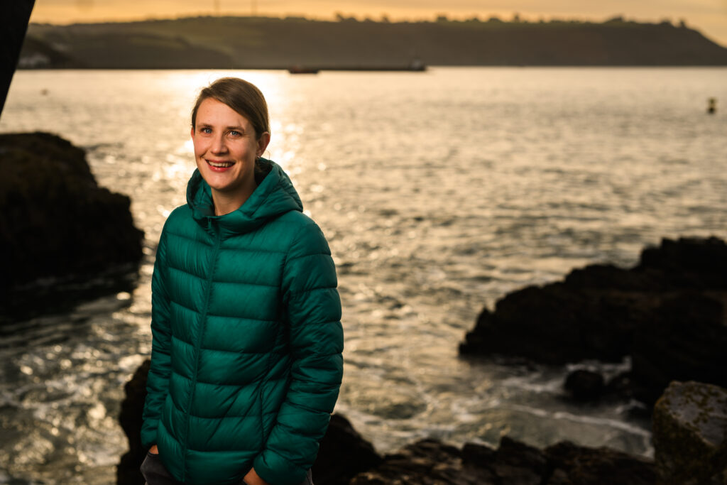 Woman with blonde hair held back, in a dark green thin puffa jacket, standing with her hands in pockets of black jeans, standing in front of the sea at sunset, with rocks and a land mass in the background.