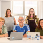 Two women stand behind three people seated at a white desk, one man and two women, with two laptops open in front of them and a large window behind the group. All are smiling at the camera.