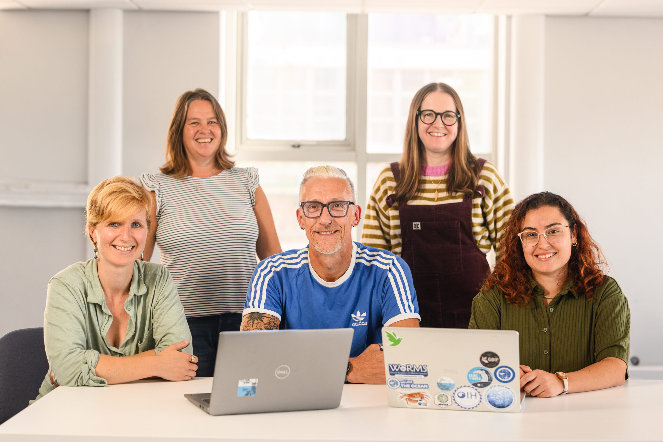 Two women stand behind three people seated at a white desk, one man and two women, with two laptops open in front of them and a large window behind the group. All are smiling at the camera.